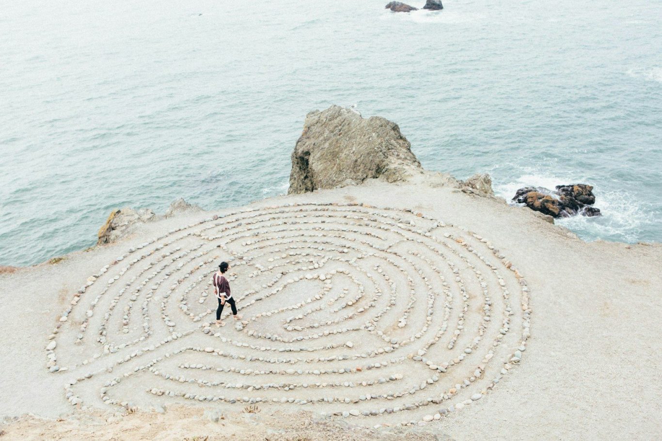 Vogelperspektive auf einen Strand mit Felsen, im Sand ein gezeichnetes Labyrinth, in das ein Mensch eintritt – Symbol für Orientierung, Refl
